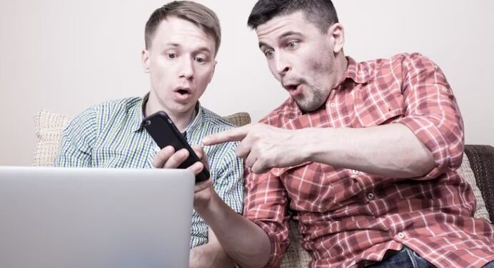 Two surprised men looking at smartphone and laptop, reacting to unexpected online content at home
